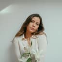 A women posing for a photo with long light brown hair and a white blouse.  She's holding white lilies.