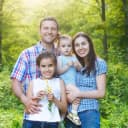 A family photo with a mother, father and two daughters set with lots of green trees in the background.