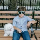 A bichon frise dog sitting with a little boy wearing sunglasses on a bench outside.
